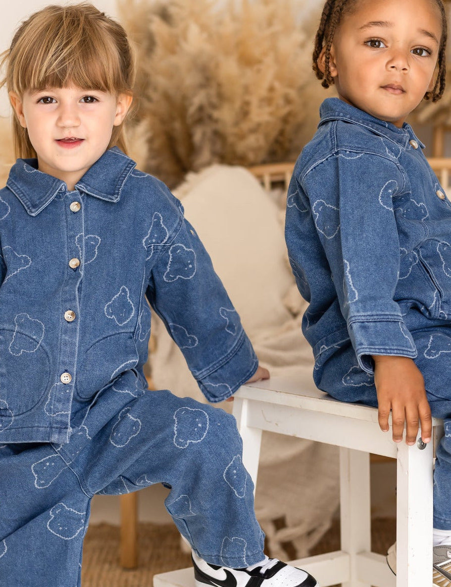 Two children wearing matching denim outfits sitting on a small white stool.