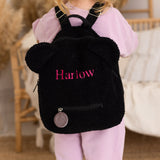 Child wearing a black backpack with 'Harlow' embroidered on it, standing indoors.