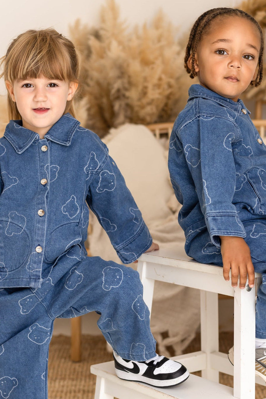 Two children wearing matching denim outfits sitting on a small white stool.