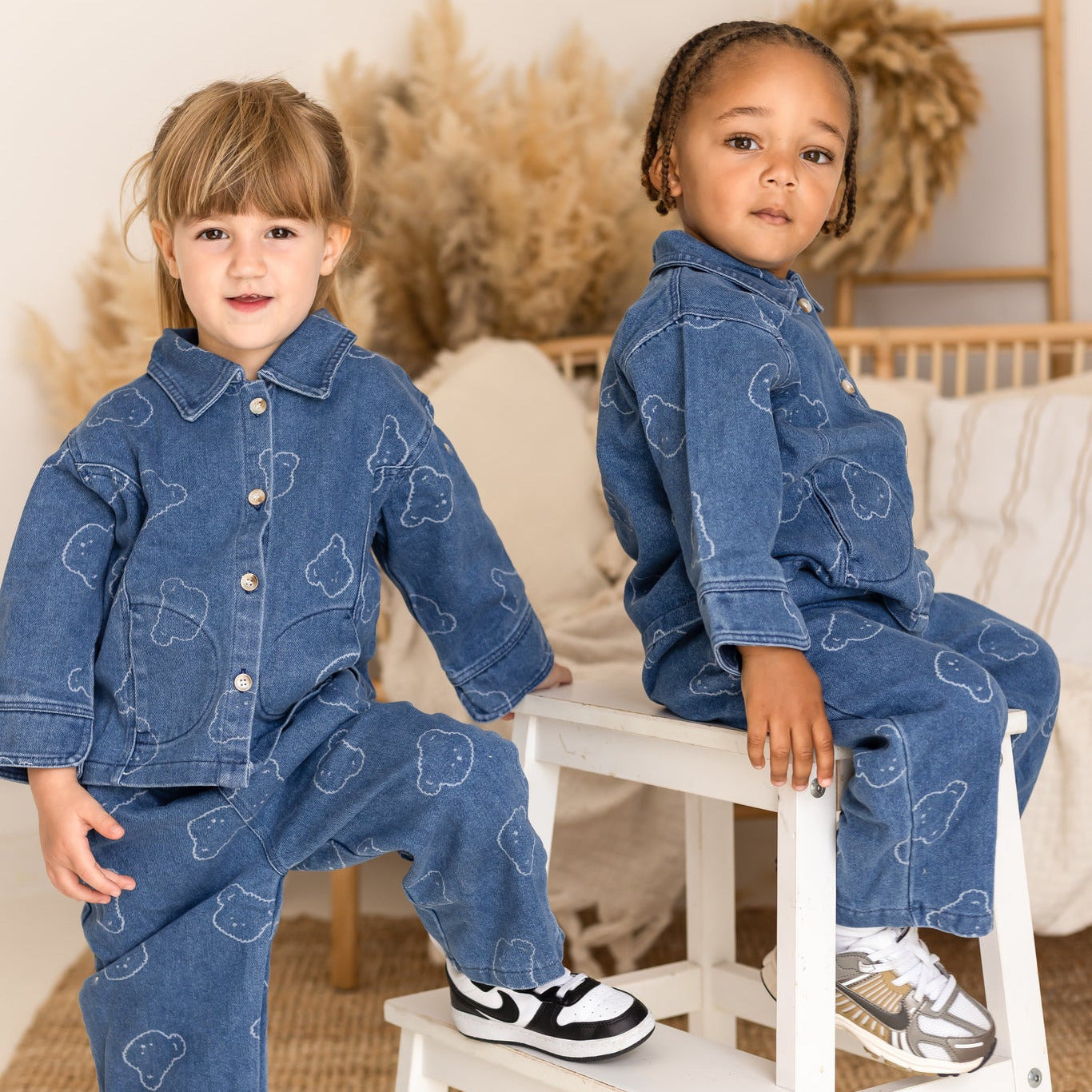 Two children wearing matching denim outfits sitting on a small white stool.