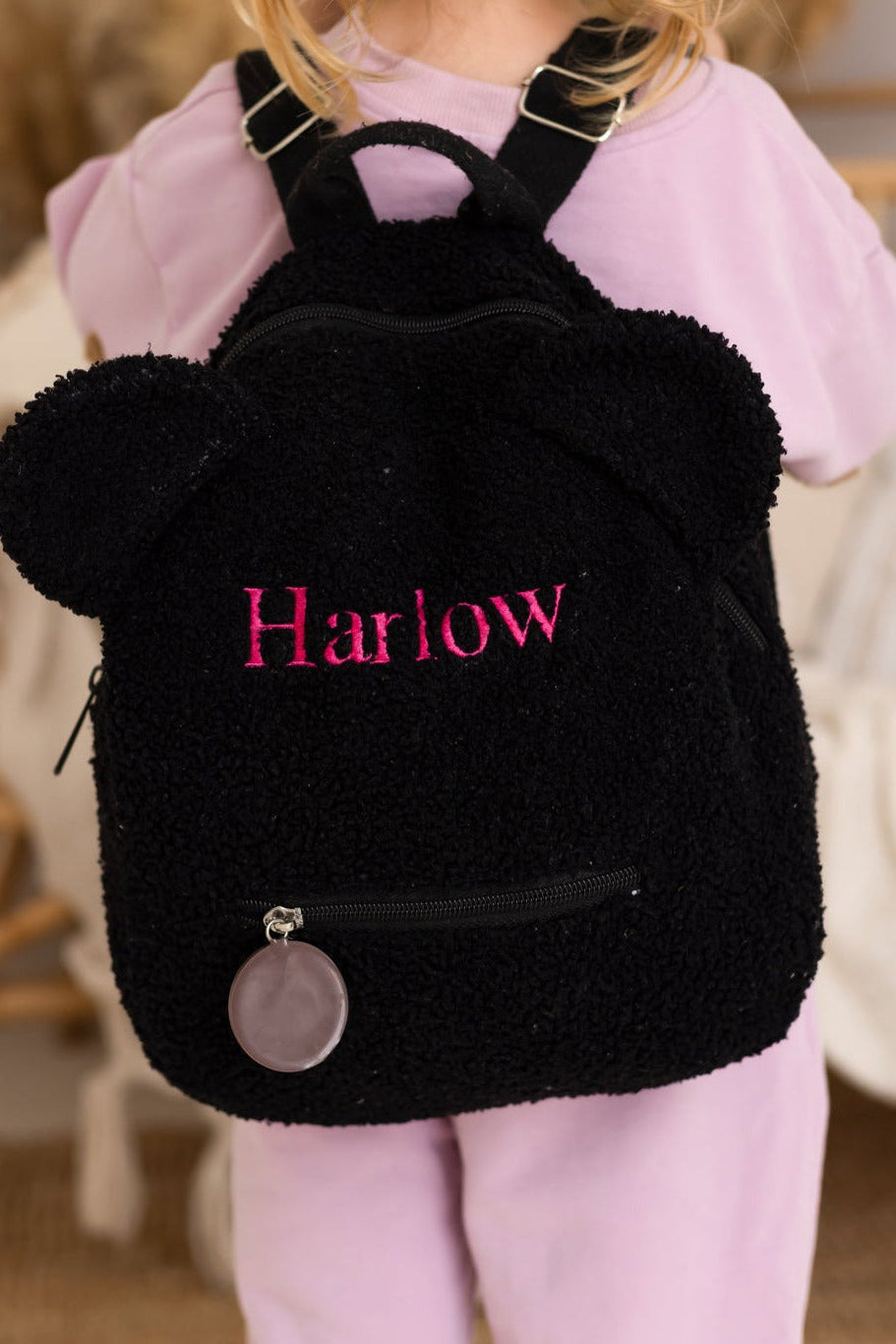 Child wearing a black backpack with 'Harlow' embroidered on it, standing indoors.