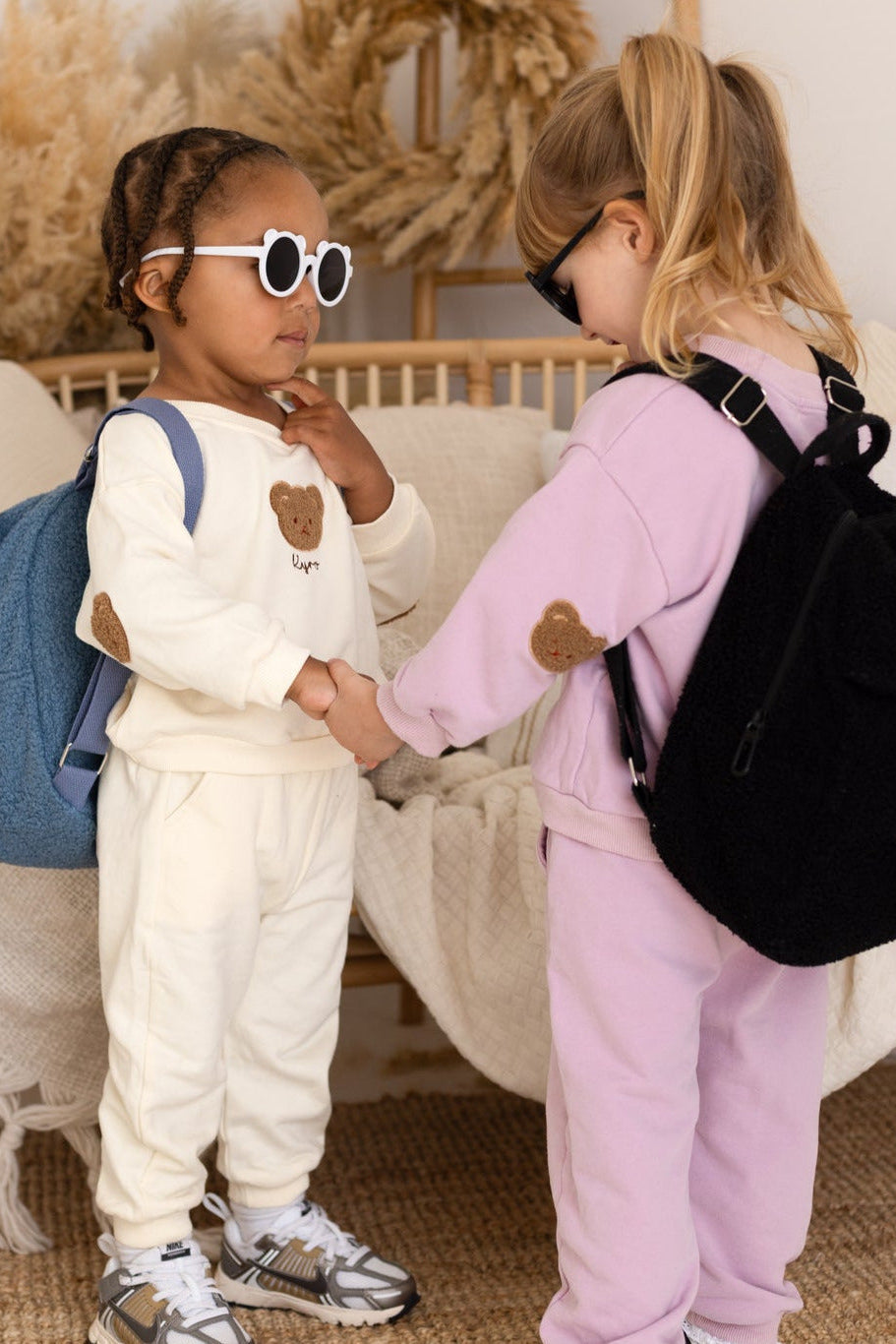 Two children in matching outfits with backpacks shaking hands in a cozy room.