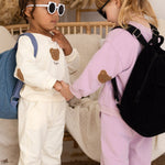 Two children in matching outfits with backpacks shaking hands in a cozy room.  The Bear Backpack Bag Personalised Cozy Crew Club Bags mora-uk.myshopify.com the-bear-backpack-bag   10off, 20off, acc, accessories, accessory, allproducts, baby, back pack, backpack, bag, beige, BFCM-20, BFCM-25, BFCM20, birthday, china, CHRISTMAS20, clothing, daywear, embroidery, kids, LINECARD, lunch box, new in, PADDY10, personalised, school, set, slip on, whatnot, WINTER20, zero